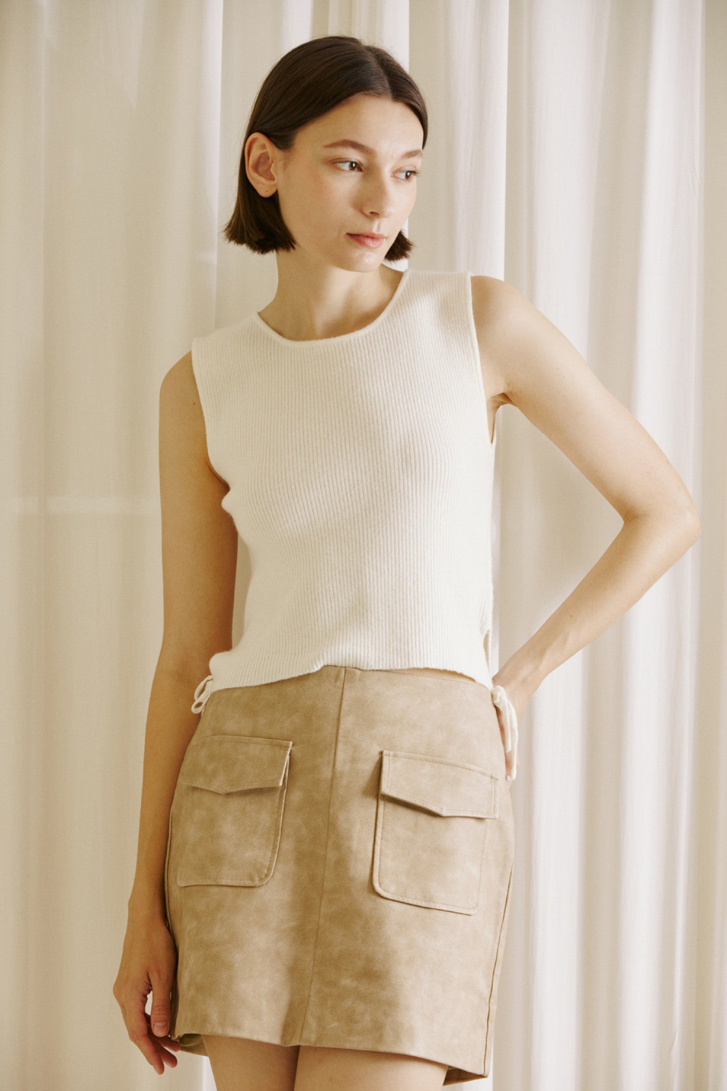 Model wearing a beige faux suede mini skirt with oversized front pockets, paired with a sleeveless cream knit top. The skirt is styled in a minimalist, neutral-tone setting, ideal for a chic fall outfit.