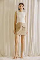 Model wearing a beige faux suede mini skirt with oversized front pockets, paired with a sleeveless cream knit top. The skirt is styled in a minimalist, neutral-tone setting, ideal for a chic fall outfit.