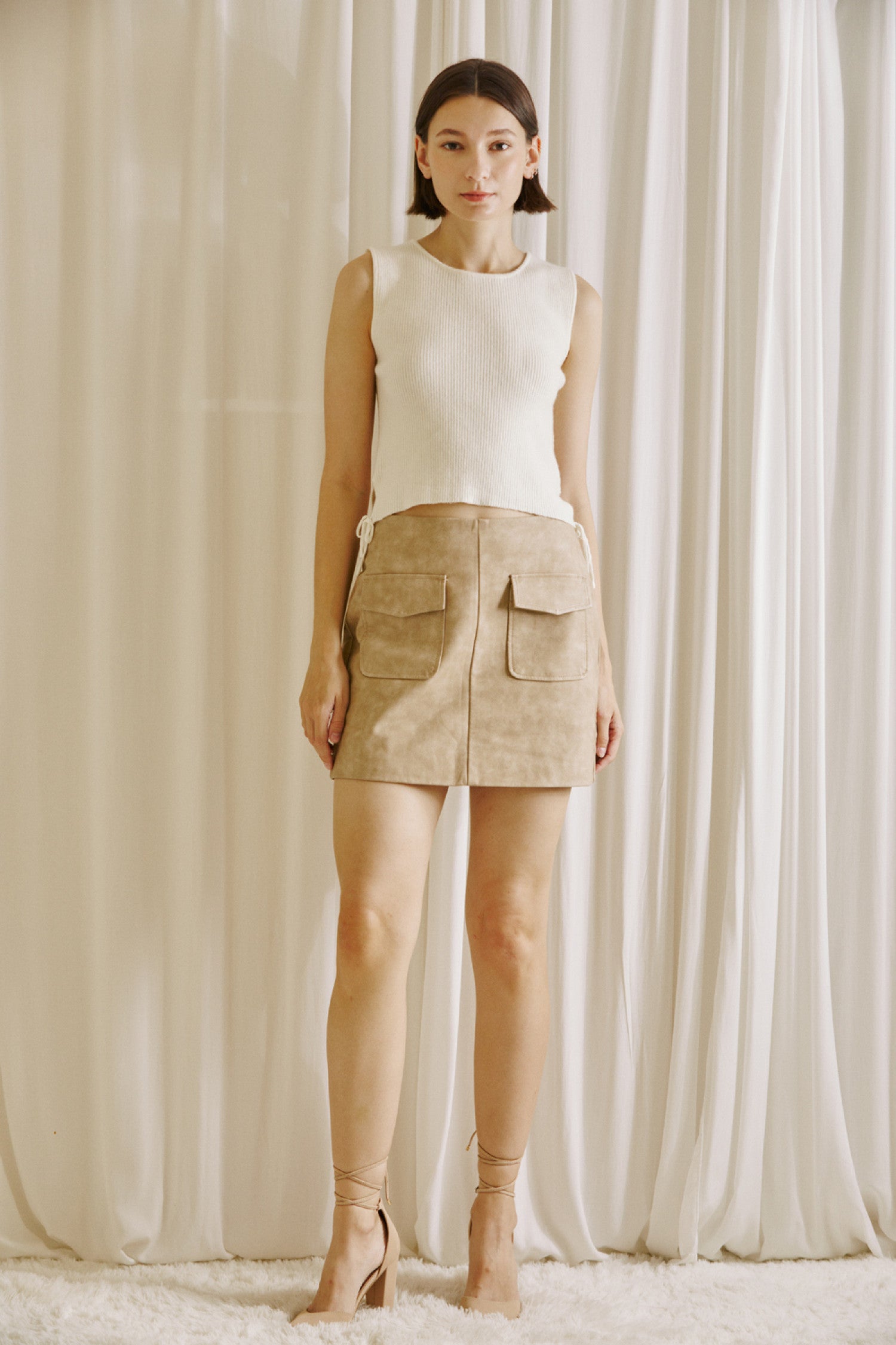 Model wearing a beige faux suede mini skirt with oversized front pockets, paired with a sleeveless cream knit top. The skirt is styled in a minimalist, neutral-tone setting, ideal for a chic fall outfit.