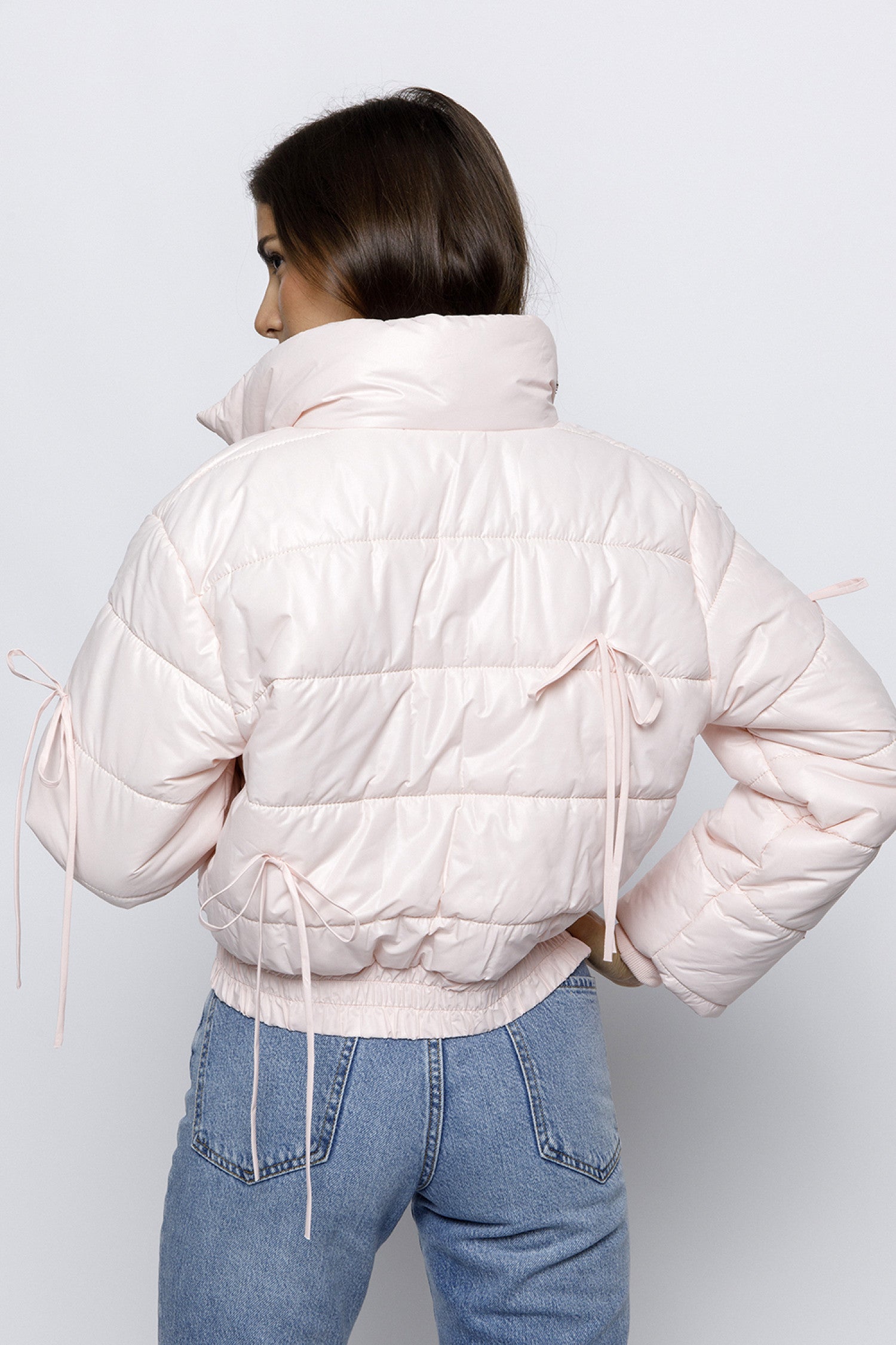 Woman wearing a light pink puffer jacket with bow details on the front and sleeves. The jacket has a high collar and quilted design, giving it a cozy and stylish look.