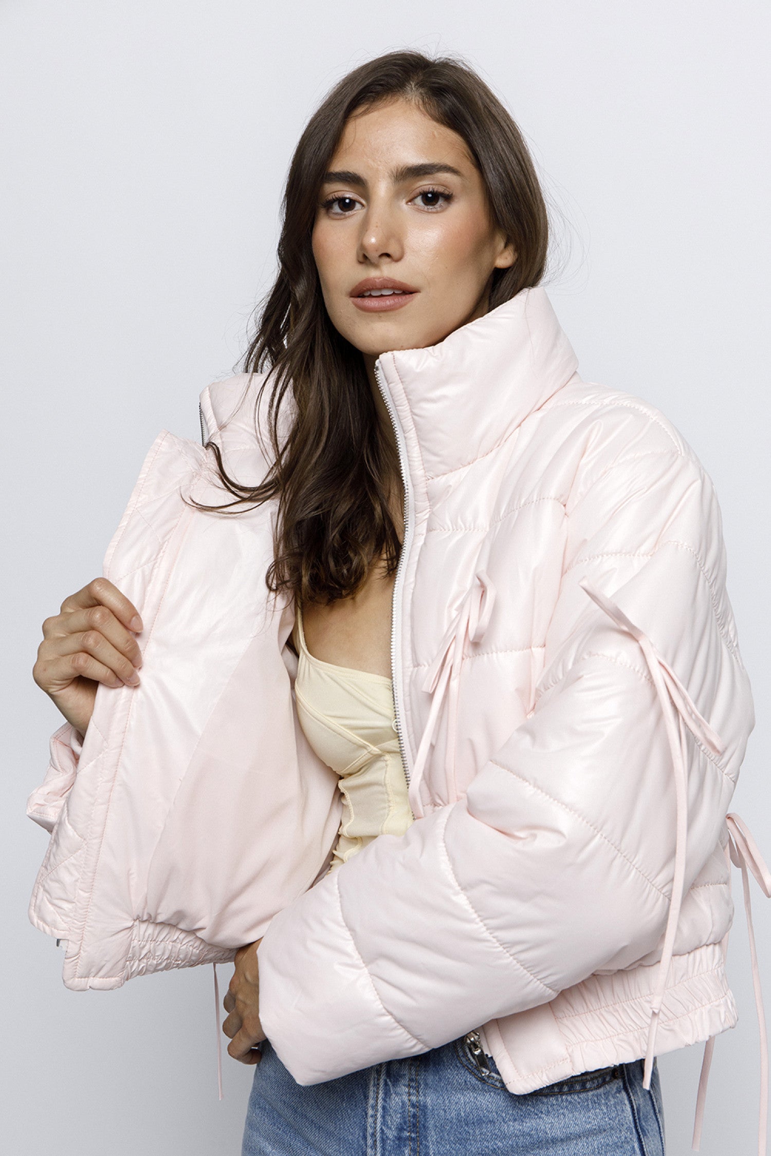 Woman wearing a light pink puffer jacket with bow details on the front and sleeves. The jacket has a high collar and quilted design, giving it a cozy and stylish look. She pairs it with light blue jeans for a casual, trendy outfit.