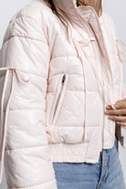 Woman wearing a light pink puffer jacket with bow details on the front and sleeves. The jacket has a high collar and quilted design, giving it a cozy and stylish look..