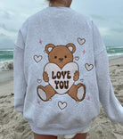 Woman wearing a cozy gray Teddy Bear Embroidered  sweatshirt by Sunkissed Coconut, featuring adorable teddy bear graphics holding "Love You" hearts on the front and a large teddy bear graphic on the back. The sweatshirt includes cute heart and star accents, perfect for a trendy, casual look on a beach day.