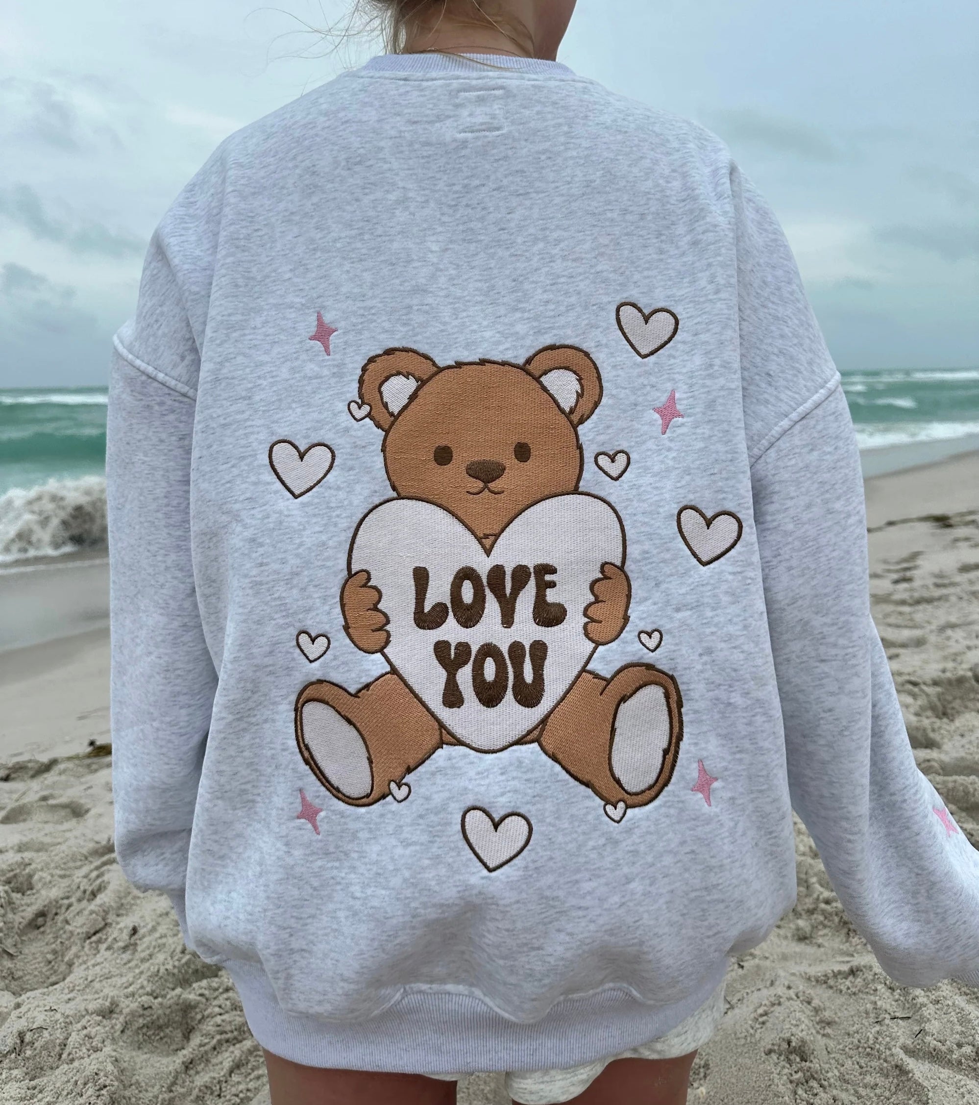 Woman wearing a cozy gray Teddy Bear Embroidered  sweatshirt by Sunkissed Coconut, featuring adorable teddy bear graphics holding "Love You" hearts on the front and a large teddy bear graphic on the back. The sweatshirt includes cute heart and star accents, perfect for a trendy, casual look on a beach day.