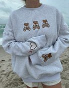 Woman wearing a cozy gray Teddy Bear Embroidered  sweatshirt by Sunkissed Coconut, featuring adorable teddy bear graphics holding "Love You" hearts on the front and a large teddy bear graphic on the back. The sweatshirt includes cute heart and star accents, perfect for a trendy, casual look on a beach day.